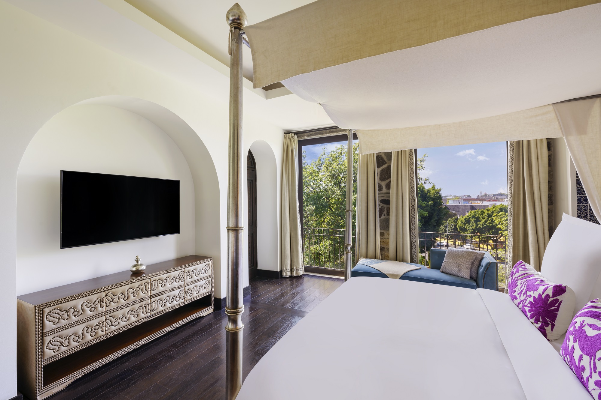 Four-poster bed facing TV in sunlit Banyan Tree Puebla Harmony suite, overlooking lush garden views.